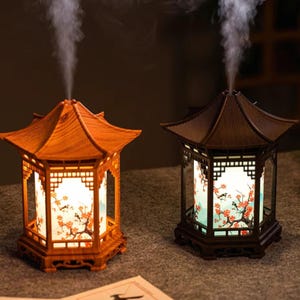Könnte beinhalten: Zwei dekorative Holzlaternen mit Dächern im Pagodenstil. Die Laternen haben ein Blumendesign auf den beleuchteten Paneelen und geben einen Dampfstrahl ab. Eine ist hellbraun, die andere dunkelbraun.