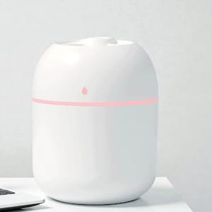 Op de afbeelding: Een witte, ovaalvormige luchtbevochtiger met een roze verlichte band en een roze waterdruppel-icoon. De luchtbevochtiger staat op een wit oppervlak, met een laptop zichtbaar in de linkerbenedenhoek. Minimalistisch en modern design.