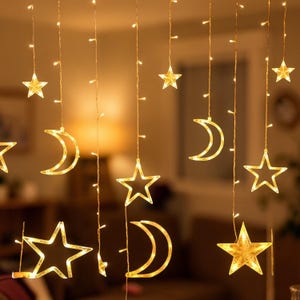 Puede incluir: Guirnalda de luces decorativa con formas de estrella y media luna, iluminada por LED blanco cálido. Las luces cuelgan de finos cables, creando un efecto celestial. Las luces tienen un suave brillo amarillo.
