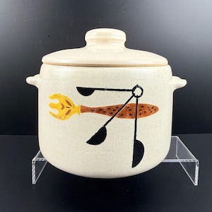 May include: Cream-colored ceramic canister with lid. The canister features a whimsical design of a yellow fork, a brown spoon, and two black utensils. The canister has two small handles and is suitable for kitchen storage.