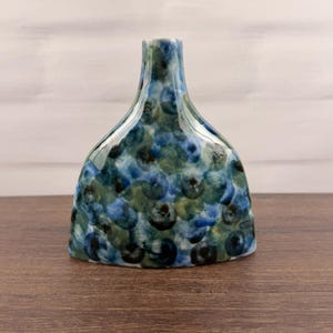 May include: A ceramic vase with a flattened, wide-bottomed shape and a narrow neck. The surface features a glossy, mottled pattern of overlapping circular shapes in various shades of deep blue, light blue, and muted green throughout the body.
