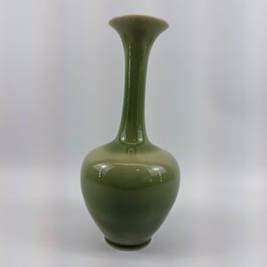 Könnte beinhalten: Eine hohe, schlanke Keramikvase mit einer glatten, glänzenden Oberfläche in einem gedämpften Olivgrün. Die Vase hat einen bauchigen, abgerundeten Boden, der sich zu einem langen, schmalen Hals verjüngt und in einer leicht ausgestellten Öffnung endet.