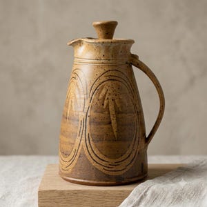 May include: A tan ceramic pitcher with a speckled finish and a matching lid. The body features two large, embossed oval patterns with leaf-like designs inside. It has a curved handle on the right side and a small pouring spout at the top rim.