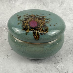 May include: A round ceramic lidded jar with a glossy light blue-green glaze and a fine crackle finish. The lid features a central circular magenta accent surrounded by a dark brown and gold crystalline pattern that radiates outward across the surface.