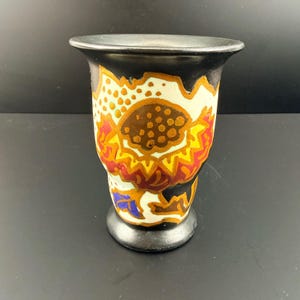 May include: A decorative ceramic vase with a black rim and base. The vase features a colorful floral design with shades of brown, yellow, red, and blue against a white background. The vase has a cylindrical shape with a slightly flared top.