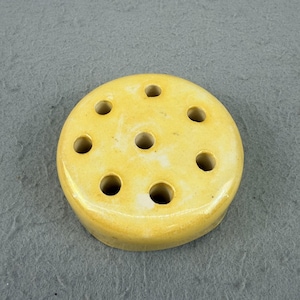 May include: A round, yellow ceramic flower frog with multiple holes. The flower arranging tool has a glazed finish and is designed to hold flower stems in place. The holes are evenly spaced across the top surface.