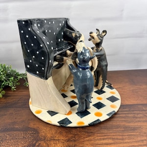 May include: A ceramic sculpture featuring a black piano with white polka dots and a cream-colored base. Two anthropomorphic dog figures are positioned at the piano, one playing the keys and the other standing, set on a round base with a diamond pattern.