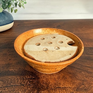 May include: A ceramic flower frog bowl with a speckled amber-orange exterior and a flat, cream-colored circular insert featuring seven evenly spaced holes for stems and a small semi-circular cutout along the edge of the insert piece.