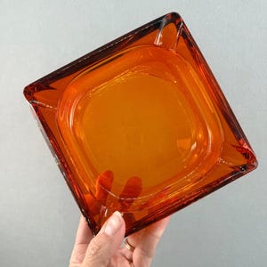 Può includere: Un posacenere quadrato in vetro color ambra. Il posacenere ha un interno quadrato arrotondato e un bordo leggermente rialzato. Il vetro è trasparente, lasciando passare la luce. Il posacenere è tenuto da una mano.