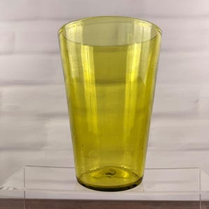 May include: A tall, tapered drinking glass made of translucent lime-yellow glass. The vessel features a smooth, cylindrical body that widens slightly toward the top rim, with a thick, solid base and subtle air bubbles embedded within the glass material.