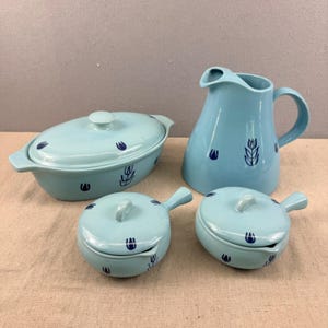 May include: A collection of light blue ceramic kitchenware featuring dark blue floral patterns. The set comprises a covered oval casserole dish, a jug, and two small lidded bowls with handles. The items are displayed on a neutral-toned surface.