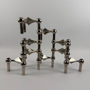 May include: A modular silver-toned metal candle holder featuring a series of interconnected spherical joints and angular, diamond-shaped connectors. The structure is composed of multiple vertical pillars of varying heights, each topped with a circular opening for a candle.