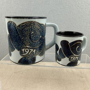 Può includere: Due tazze in ceramica con base bianca e design astratto blu. Le tazze hanno un bordo nero e l'anno "1971" stampato sul davanti. Una tazza è più grande dell'altra, con un manico azzurro.