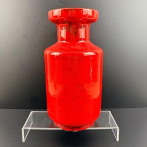 Könnte beinhalten: Eine leuchtend rote Keramikvase mit glänzendem Finish. Die Vase hat einen zylindrischen Körper, einen ausgestellten Hals und einen breiten, flachen Rand. Die Vase wird auf einem transparenten Acrylregal vor dunklem Hintergrund ausgestellt.