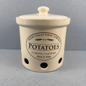 Puede incluir: Recipiente para patatas de cerámica color crema con tapa y pomo. El recipiente tiene el texto "POTATOES" en un diseño circular con "FRESH VALLEY FARM COMPANY" y "ST. HELENA, CALIFORNIA SINCE 1918". Tiene tres agujeros circulares cerca de la base.