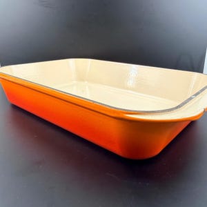 May include: A rectangular orange baking dish with a cream interior. The dish has a glossy finish and is made of a durable material. The sides slope outwards, and the dish appears unused. The dish measures approximately 30 cm in length.