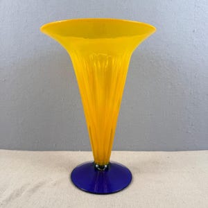 Puede incluir: Un jarrón alto de vidrio amarillo con una parte superior acampanada y un diseño acanalado. El jarrón tiene una base circular azul. El jarrón mide aproximadamente 25 cm de alto.