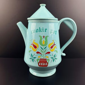 Può includere: Una teiera per biscotti in ceramica azzurra con coperchio e manico. La teiera presenta un motivo floreale con accenti rossi, gialli e verdi, e le parole "cookie pot" sono stampate sul davanti.