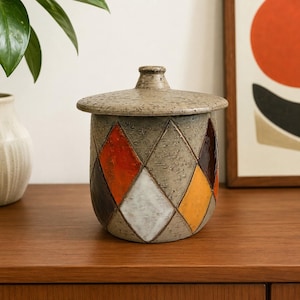 May include: A cylindrical ceramic jar with a matching lid, featuring a textured gray surface decorated with a repeating diamond pattern in shades of orange, yellow, white, and dark brown. The jar has a slightly tapered base and a small knob handle.