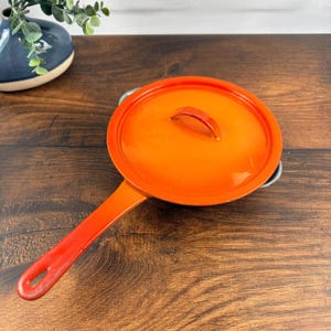 Op de afbeelding: Een feloranje geëmailleerde gietijzeren koekenpan met bijpassend deksel. De pan heeft een lange, rechte steel met een gat aan het uiteinde om op te hangen en een gebogen handvat aan de tegenoverliggende zijde. Het deksel heeft een boogvormige greep.