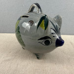 Może przedstawiać: Szara ceramiczna skarbonka z uchwytem i malowanymi detalami. Bank posiada fantazyjny wzór z niebieskim pyskiem, okiem oraz zielonymi i żółtymi akcentami liści. Bank ma trzy małe nóżki.
