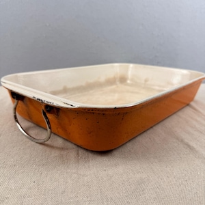 May include: Orange rectangular baking dish with white interior and metal handles. The enamel-coated dish has a distressed appearance, with visible wear and tear. The dish is empty and ready for use.