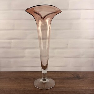 Pode incluir: Um vaso de vidro alto e esguio com um tom suave de malva ou rosa empoeirado. O vaso apresenta uma abertura larga e alargada em forma de trombeta no topo que se estreita para um pescoço fino, apoiado numa base circular transparente.