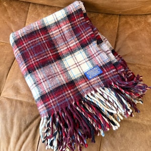 Puede incluir: Una manta a cuadros en tonos rojos, burdeos, crema y azul. La manta tiene un borde con flecos y una etiqueta azul con texto blanco. La manta está doblada y sobre una superficie marrón.