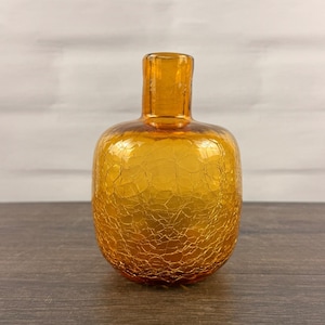 Pode incluir: Um pequeno vaso de vidro redondo em uma vibrante cor laranja âmbar. A superfície do vaso apresenta uma textura craquelada distinta em todo o corpo, com um pescoço liso e estreito e uma abertura circular no topo para hastes de flores.