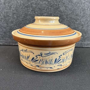 May include: A ceramic lidded casserole dish. The lid is tan with brown and blue stripes. The dish is tan with a brown rim and a blue and brown grass design around the base. The dish is round.