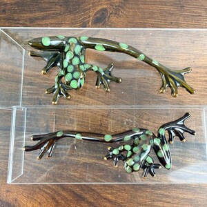 May include: Two dark brown ceramic frog-shaped drawer pulls with light green spots. Each handle features a long, slender body and four splayed feet with gold-toned edges, designed for mounting onto furniture surfaces with a horizontal orientation.