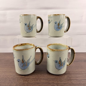 May include: A set of four ceramic mugs with a speckled light gray finish and a brown rim. Each mug features a hand-painted blue bird with orange wing details and a small butterfly. The mugs have a cylindrical shape and a curved handle on the side.