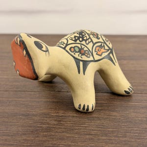 Puede incluir: Una figura de tortuga de cerámica de color crema con una cara plana de color marrón rojizo y un caparazón decorado con segmentos geométricos delineados en negro que contienen pequeñas ilustraciones pintadas de insectos y animales.