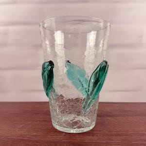 May include: A clear glass tumbler featuring a distinct crackle texture throughout the body. The vessel is adorned with three applied decorative glass elements in a vibrant teal green color, each shaped like a smooth, elongated leaf or petal.