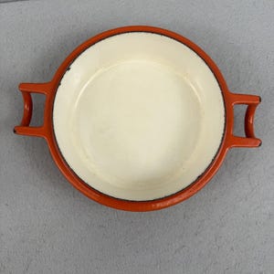 May include: Round, orange and cream-coloured cast iron dish with two handles. The dish has a cream-coloured interior and an orange exterior with a black rim. The handles are also orange.