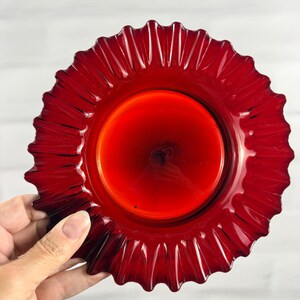 Könnte beinhalten: Ein runder, durchscheinender Teller aus rubinrotem Glas mit einem tief gerüschten, gewellten Rand. Die Mitte des Tellers ist glatt und flach und geht in eine Reihe von strahlenförmigen, vertikalen Rippen über, die zum gezackten Rand führen.
