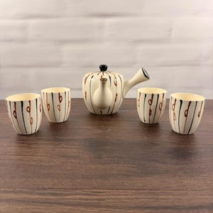 Puede incluir: Un juego de té de cerámica que consta de una tetera con asa lateral y cuatro tazas a juego. Cada pieza presenta una base de color crema con una superficie texturizada, decorada con líneas verticales negras y pequeños detalles ovalados de color marrón.