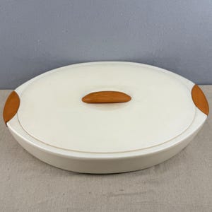 May include: Cream-coloured oval serving dish with a matching lid. The lid has a wooden handle and wooden accents on each end. The dish is designed for serving food and has a simple, elegant design.