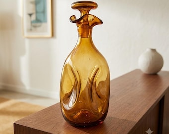 May include: A translucent amber-colored glass decanter featuring a rounded body with indented sides, a narrow neck, and a flared, ruffled rim with a matching glass stopper. The glass contains small air bubbles throughout its structure.
