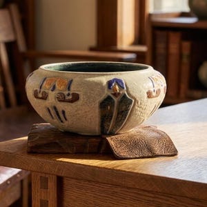 May include: A round ceramic bowl with a textured off-white exterior featuring embossed geometric patterns in blue, brown, and gold. The interior of the bowl is glazed in a deep green color. The vessel has a wide, low-profile shape with a smooth rim.