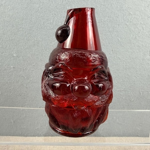 Könnte beinhalten: Ein tiefrotes, durchscheinendes Glasgefäß in Form einer Weihnachtsmannfigur. Das Gefäß zeigt ein geformtes Gesicht mit einer markanten Nase, einem Schnurrbart und einem Bart, gekrönt von einer spitzen Mütze mit einem kleinen runden Bommel.