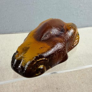 Puede incluir: Un objeto de vidrio marrón ámbar, posiblemente una pieza decorativa o una escultura. El objeto tiene una forma lisa y redondeada con una forma ligeramente irregular. El vidrio parece translúcido, con variaciones de color y tono.