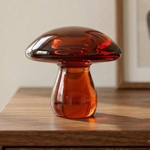 Pode incluir: Uma figura de cogumelo em vidro âmbar translúcido com um chapéu largo e arredondado e um caule cilíndrico espesso. O objeto é feito de vidro liso e polido que reflete a luz, criando gradientes de cores profundas em tons de laranja e castanho.