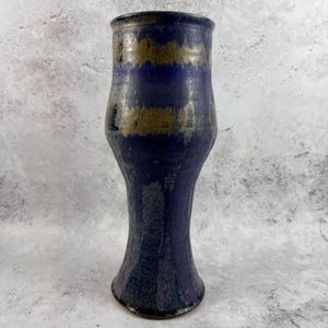 May include: A tall, cylindrical ceramic vase featuring a deep blue glaze with horizontal bands of muted gold and bronze tones. The surface has a textured, mottled appearance with a slightly flared base and a smooth, rounded rim at the top.
