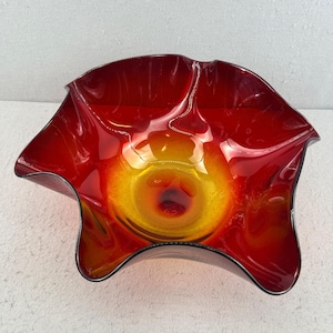 Puede incluir: Un cuenco decorativo de vidrio con un gradiente de color vibrante. El cuenco pasa de un rojo intenso en los bordes a un centro amarillo brillante, con un centro rojo oscuro. El borde tiene una forma ondulada y orgánica y un contorno negro.