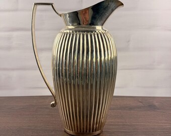 May include: A silver-toned metal pitcher featuring a prominent vertical ribbed texture along its rounded body. The vessel includes a high, arched handle attached to the side and a wide, smooth pouring spout at the top rim, creating a balanced silhouette.