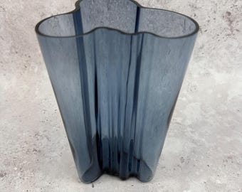 May include: A blue glass vase with a wavy rim and a flared, textured body. The vase has a unique, organic shape and a translucent quality, suitable for displaying flowers or as a decorative piece. The vase is approximately 10 inches tall.