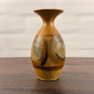 Puede incluir: Un pequeño jarrón de cerámica bulboso con borde acampanado, que presenta un acabado moteado en tonos bronceado y marrón. El cuerpo está decorado con dos prominentes patrones circulares de pinceladas marrón oscuro y bandas horizontales.
