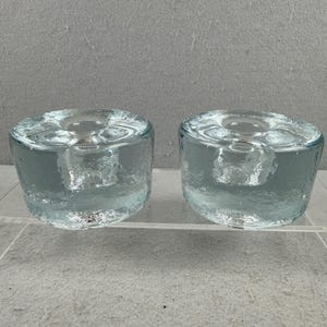 Puede incluir: Dos candelabros cilíndricos de vidrio transparente. Cada soporte tiene una superficie texturizada y una hendidura circular en la parte superior para una vela. El vidrio tiene un ligero tinte azul-verdoso, y los soportes se exhiben sobre una superficie transparente.