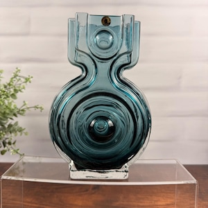 Puede incluir: Un jarrón de cristal azul cerceta con un cuerpo circular con patrones de ondas concéntricas y una sección superior geométrica escalonada. Una pequeña pegatina circular es visible en el borde superior de la estructura de cristal.